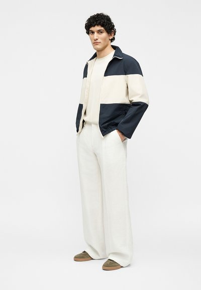 Νεαρός άνδρας στέκεται με τα χέρια στις τσέπες, φορώντας ένα μπουφάν σε ναυτικό μπλε και λευκό, μπεζ πουλόβερ, φαρδιά λευκά παντελόνια και καφέ αθλητικά παπούτσια.