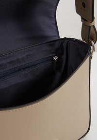 Beige olkalaukku, jossa on vetoketjulla suljettava sisätasku, vuorattu mustalla kankaalla. Sujuva pinta ja virtaviivainen muotoilu säädettävällä hihnalla.