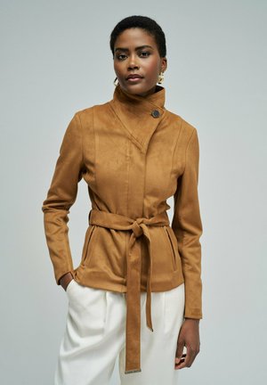 Femme portant une veste marron ceinturée avec un col montant et un pantalon blanc, debout avec une main dans la poche, sur un fond uni.