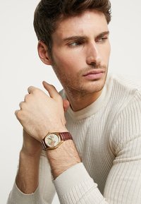 Round gold watch with a textured cream dial and a burgundy leather strap, featuring a detailed illustration on the face.