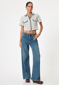 Kvinde iført en hvid cropped skjorte med knapper og sort kant, højtaljede vide blå jeans og brune platformstøvler foran en hvid baggrund.