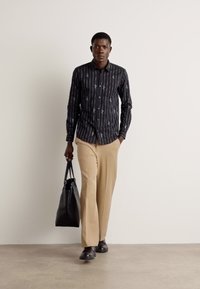 Camicia nera con bottoni, a righe verticali e motivi astratti, abbinata a pantaloni beige, borsa a tracolla nera e scarpe nere.