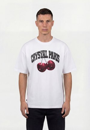 Mann trägt ein weißes T-Shirt mit roten Kirschen und dem Schriftzug „Crystal Paris“, steht vor einem einfarbigen hellen Hintergrund.
