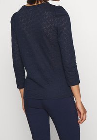 Námořnicky modrý, půlprůhledný top s texturovaným vzorem, 3/4 rukávy a zaoblený výstřih. Má jemné nadýchané detaily na ramenou.