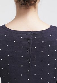 Granatowy sweter z dzianiny z białym kropek wzorem, okrągłym dekoltem i pięcioma małymi guzikami z tyłu. Gładka tekstura z delikatnym prążkowaniem.
