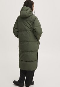 Langer, voluminöser, olivgrüner Mantel mit Kapuze, der über ein gestepptes Design, seitliche Schlitze und eine glatte Textur verfügt. Sichtbare schwarze Hose und klobige schwarze Stiefel.