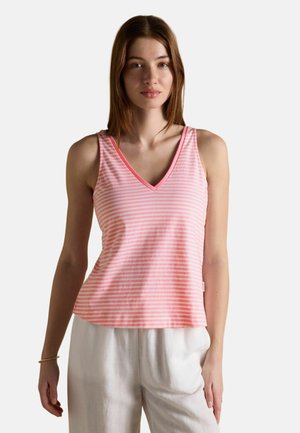 Młoda kobieta w różowo-białym, paskowanym topie bez rękawów z dekoltem w kształcie litery V i białych spodniach, stojąca z neutralnym wyrazem twarzy na jednolitym tle.