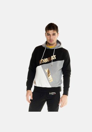 Giovane uomo che indossa una felpa con cappuccio color-block nera, grigia e bianca con scritte dorate, in piedi davanti a uno sfondo chiaro uniforme.