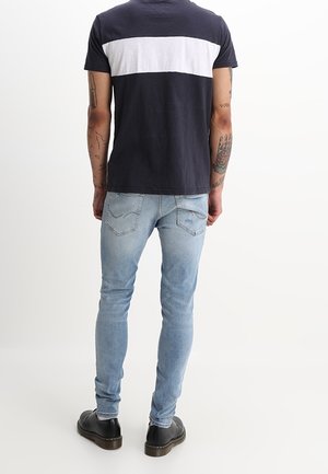 T-shirt bleu foncé avec une bande horizontale blanche, associé à un jean skinny bleu clair et des chaussures à lacets noires, vu de dos.