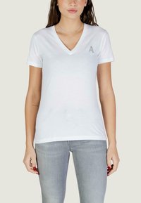 Femme portant un t-shirt blanc à col en V avec un petit emblème argenté en forme de « A » sur la poitrine gauche, associé à un jean gris clair, posant devant un fond uni.