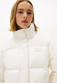 Chaqueta blanca acolchada con un cuello alto, cierre de cremallera y un pequeño parche de la marca en el lado izquierdo. Textura suave con un patrón acolchado.