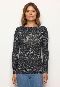Eine Frau trägt ein enganliegendes Longsleeve-Oberteil mit schwarzem und grauem Leopardenmuster sowie eine blaue Jeans und steht vor einem schlichten weißen Hintergrund.