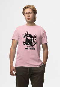 Růžové bavlněné tričko s černým abstraktním designem sedící postavy a textem "Matisse" ve spodní části. Kulatý výstřih, krátké rukávy.