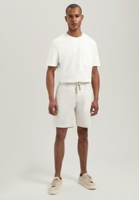 Weißes Baumwoll-T-Shirt mit einer Brusttasche, kombiniert mit hellbeigen Shorts mit Kordelzug. Braune Sneakers runden das Outfit ab. Glatte Textur, lässiges Design.