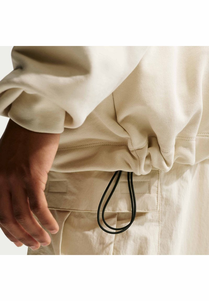 Personne portant un sweat-shirt et un pantalon beiges, main gauche détendue sur le côté, avec un cordon noir en boucle pendant à la taille.