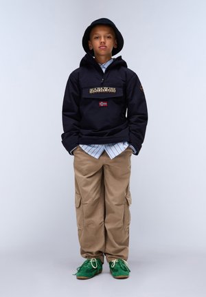 Chaqueta negra con cremallera y parche de logo, pantalones cargo caqui y zapatillas verdes. El conjunto se complementa con una camiseta de rayas azul claro debajo.