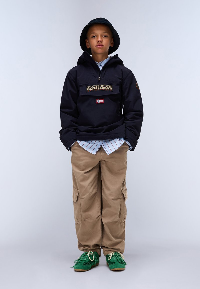 Svart genserjakke med logo-patch, khaki cargo-bukser og grønne sneakers. Utstyret er lagd med en lyseblå stripete skjorte under.