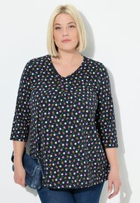 Blonde Frau in einem marineblauen Shirt mit einem Muster aus Teddybären, Fußbällen und pinken Schleifen, die eine blaue Jeans-Clutch-Tasche hält und vor einer weißen Wand steht.
