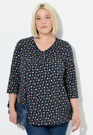 Blonde Frau in einem marineblauen Shirt mit einem Muster aus Teddybären, Fußbällen und pinken Schleifen, die eine blaue Jeans-Clutch-Tasche hält und vor einer weißen Wand steht.