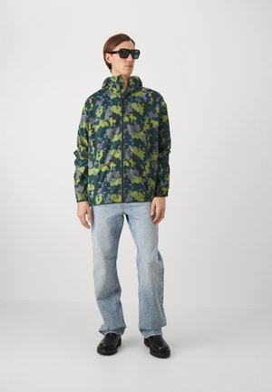 Giacca a vento con motivo mimetico in verde e blu navy, con zip frontale, cappuccio e polsini elasticizzati, abbinata a jeans blu chiaro e scarpe nere.