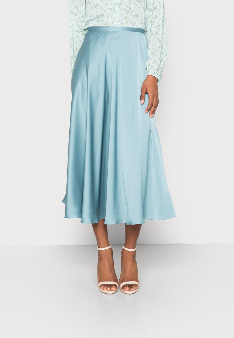 Femme portant une jupe midi en satin bleu clair, un chemisier léger à motifs floraux et des sandales à talons blanches avec bride à la cheville, debout devant un fond uni.