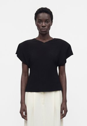 Femme aux cheveux courts portant un haut noir col en V avec des épaules larges et un pantalon plissé crème taille haute, debout devant un fond uni.