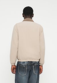 Μπεζ μπο υφάν με καφέ γιακά, ελαστικά μανίκια και ζώνη στη μέση, εμφανίζεται από πίσω. Φορεμένο με σκούρο τζιν τζιν που έχει ξεθωριασμένο μοτίβο.