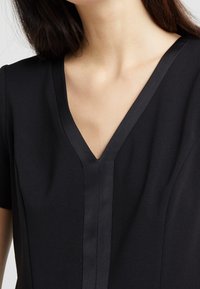 Černé šaty s výstřihem do V, krátkými rukávy a saténovým lemem kolem výstřihu a předního rozparku, vyznačující se hladkým povrchem látky.