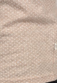 Tela beige claro con un patrón texturizado de pequeños semicírculos blancos. Incluye una etiqueta visible en el lado derecho. Textura suave.
