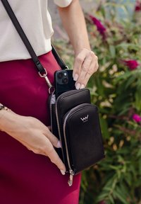 Black leather crossbody wallet with two zippered compartments, featuring silver hardware and a logo on the front, held next to a smartphone.