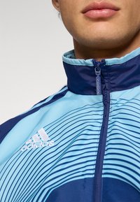 Μπλε και γαλάζιο φερμουάρ με οριζόντιες ρίγες. Διαθέτει γιακά χαμηλής στάσης και λευκό λογότυπο Adidas στο στήθος. Ομαλή υφή.