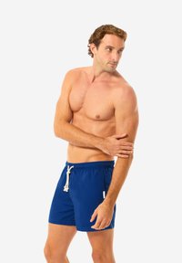 Shorts da nuoto blu realizzati in un tessuto leggero, dotati di una cintura elastica con cordino. Texture liscia, lunghezza al ginocchio, branding minimale.