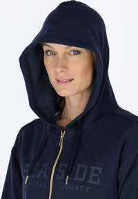 Navy zip-up hoodie with a gold zipper, large hood, and prominent "SEASIDE" text design. Made of soft fabric with a smooth texture.