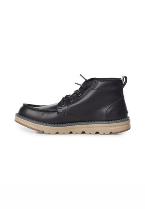Stivaletto alla caviglia in pelle nera con suola beige scanalata, chiusura frontale con lacci e tirante sul tallone, mostrato di profilo su sfondo bianco.