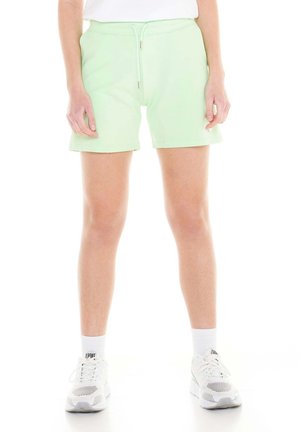 Une personne porte un short vert pâle à cordon, un haut blanc, des chaussettes blanches et des baskets blanches sur un fond blanc.