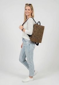 Smiling woman wearing sunglasses on her head, white sweater, light jeans, white shoes, carrying a brown geometric patterned backpack.