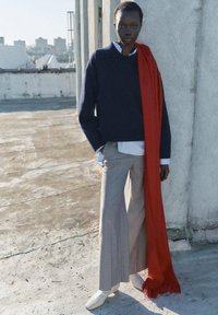 Pulover tricotat navy, cămașă albă cu guler, pantaloni largi gri și o eșarfă roșie cu ciucuri, purtate în aer liber pe o suprafață de beton.