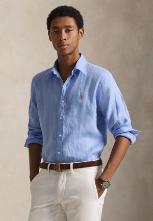 Camicia di lino azzurro chiaro con colletto button-down e piccolo logo ricamato, abbinata a pantaloni bianchi e cintura in pelle marrone.