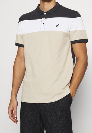 Mann trägt ein kurzärmeliges Poloshirt mit schwarzen, weißen und beigen horizontalen Streifen und dunkle, strukturierte Hosen, eine Hand in der Tasche.