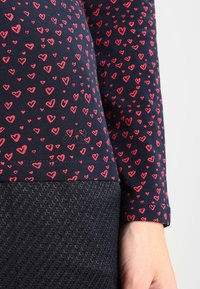 Tela azul marino con patrones de corazones rojos en las mangas y el torso, combinada con una falda texturada de un azul oscuro, y una mano visible al lado.