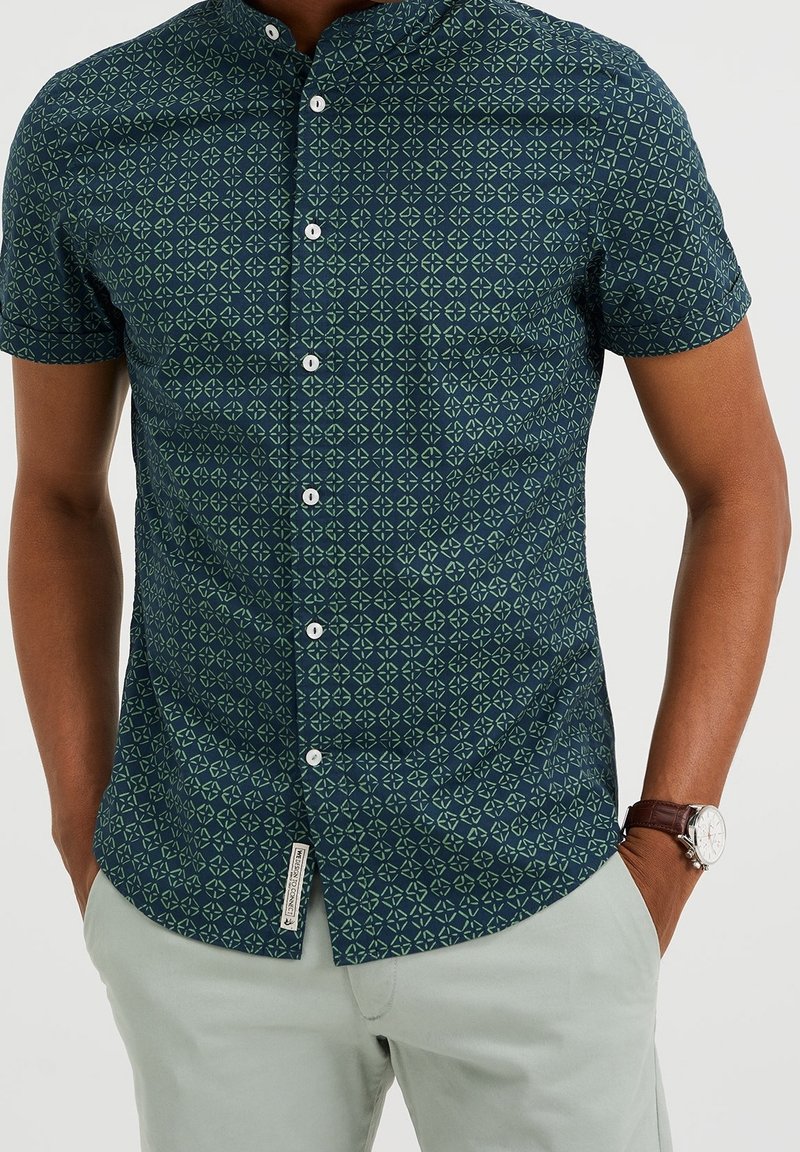 Kortärmad skjorta med knappar i mörkblått med ett geometriskt grönt mönster, med vita knappar och en liten märkeslapp vid fållen.