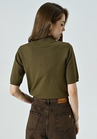 Maglione corto a maniche corte verde oliva con colletto ripiegato, abbinato a jeans a vita alta marrone scuro con tasche posteriori visibili e etichetta.