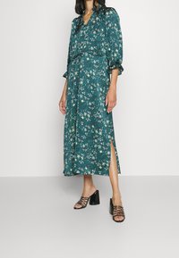 Vestido maxi floral en tejido azul verdoso con mangas abullonadas, cintura elástica, abertura lateral y un estampado de pequeñas flores blancas. Combinado con sandalias de tacón bloque negro.