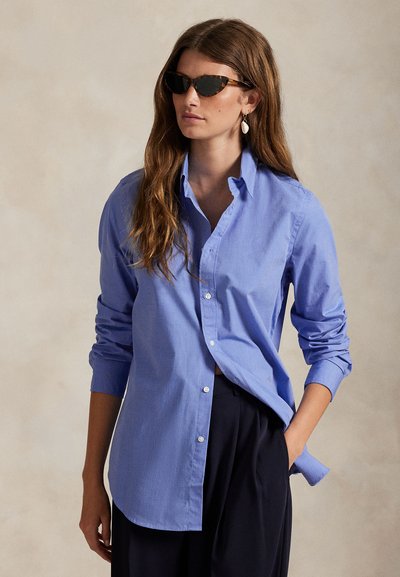 Γυναίκα με μακριά καστανά μαλλιά, φορώντας γυαλιά ηλίου με καστανόλευκη εκρού, μπλε πουκάμισο, μαύρο παντελόνι και σκουλαρίκια από κοχύλια, στέκεται με το ένα χέρι στην τσέπη.