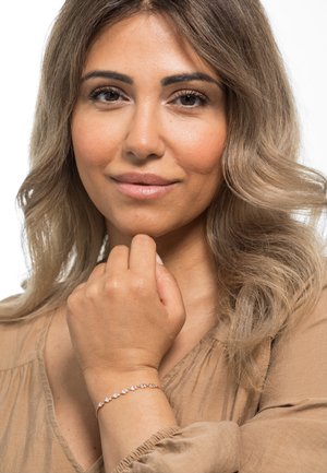 Goldarmband mit kleinen, klaren Steinen, die in einem zarten Muster angeordnet sind. Das Model hat hellbraunes Haar und eine gebräunte Bluse und zeigt das Armband an ihrem Handgelenk.