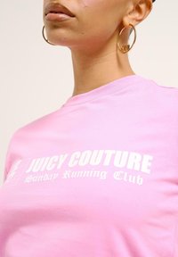 Rozā kokvilnas t-krekls ar baltu tekstu "JUICY COUTURE Sunday Running Club." Īsas piedurknes un apaļš izgriezums, ar gludu tekstūru.