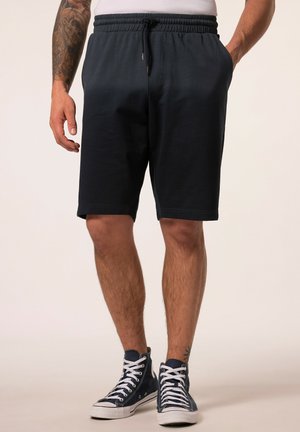 Mann trägt schwarze Shorts mit Kordelzug und dunkelblaue High-Top-Sneakers, steht mit den Händen in den Taschen.