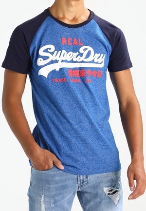 Mężczyzna ubrany w niebiesko-granatową koszulkę raglanową z logo "Real SuperDry" oraz przetarte jasnoniebieskie dżinsy, z ręką w kieszeni.