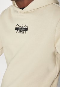 Felpa in cotone beige con cappuccio regolabile, caratterizzata da un logo "Calvin Klein" nero sul davanti. Tessuto liscio con vestibilità comoda.