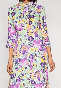 Kukallinen mekko vaaleanvihreänä purppuran, keltaisen ja vaaleanpunaisen sävyillä. Napitettava etuosa, kolmannen neljänneksen hihat ja löysä, virtaava muoto.